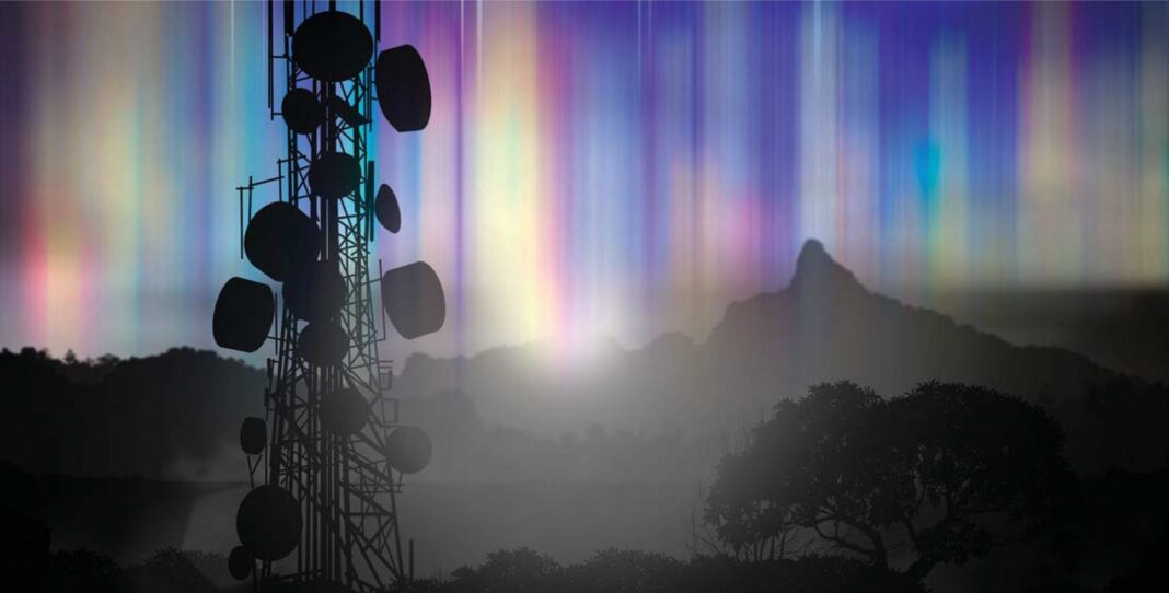 Cell tower silhouette with colorful aurora over mountains.
